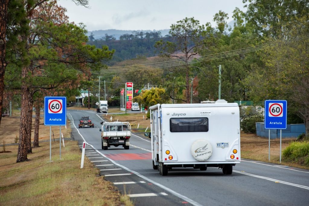 Back of Camper trailer that is driving along road in bushy rural area, through a 40 zone with petrol station ahead.