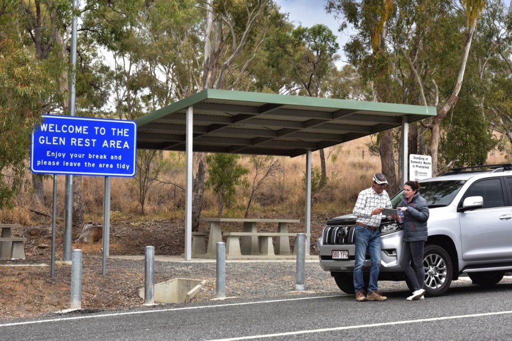 Safe Road Tripping. A man and a woman stand leaning against a silver car parked at a rest stop and look at a map.