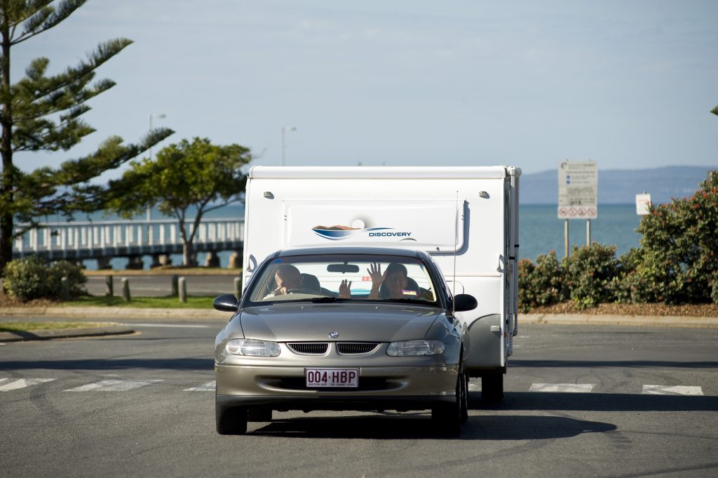 Two seniors drive off in a silver sedan towing a trailer at car park near the ocean.