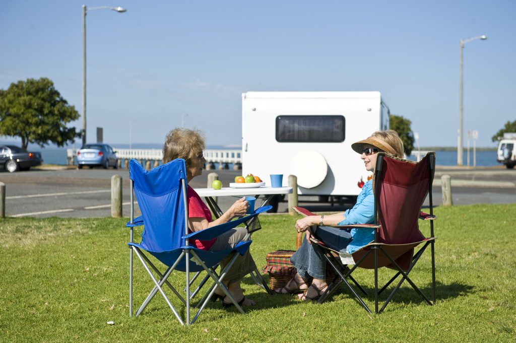 Two female RV drivers sit on camper chairs on grass outside their campervan with ocean in the background.